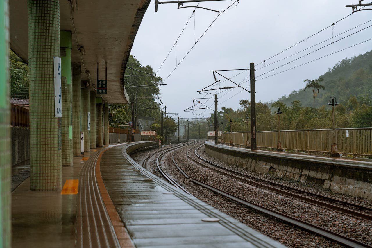 Empty train station platform in rainy weather, showcasing wet tracks and overcast skies.