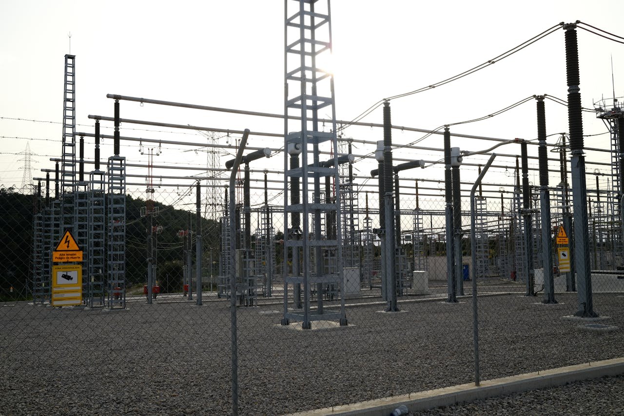 Outdoor electrical power substation with high voltage equipment and safety signs, surrounded by fencing.